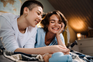 Two lesbian girls smiling and holding photo while lying in bed at home