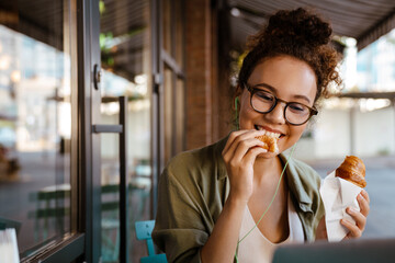 White young woman eating croissant while using laptop in cafe outdoors