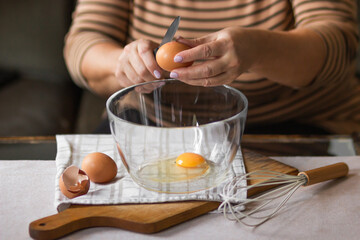 Old woman's hands cracking eggs into glass bowl with knife. Prepare for making omelet or pancakes for breakfast.