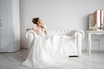 Fototapeta premium the bride in a wedding dress sits on the sofa spreading her train in a bright interior, gently touching her neck with her finger and enjoying the moment