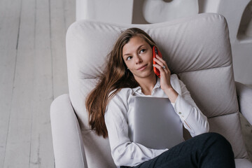 Puzzled attractive hispanic woman in white shirt laying on cozy sofa holds laptop, talks by phone at home. Beautiful Caucasian businesswoman remote working. Business and finance.