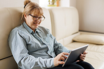 Beautiful mature woman in eyeglasses is using a digital tablet and smiling while sitting on couch at home