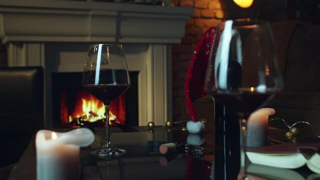 Glass of red wine on table at home at Christmas and wine bottle with Santa hat. Winter mood with fireplace in cosy dark living room.