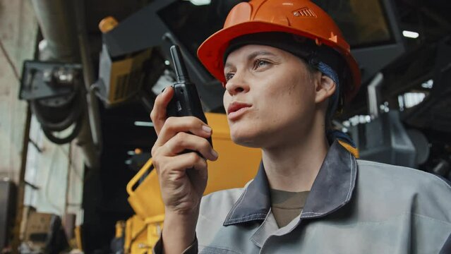 Confident female engineer in hard hat speaking to employees through walkie talkie operating work at tractor plant