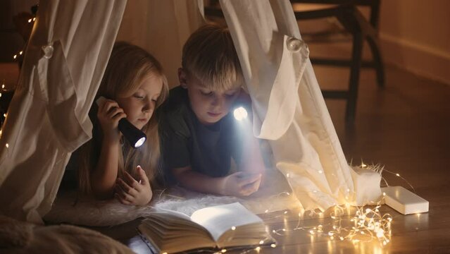 curious little children are reading book with fairytales in handmade teepee in room