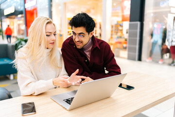 Happy young couple using typing laptop computer together, discussing purchase on the Internet, looking to screen sitting on sofa at table in hall of shopping mall with bright modern interior.