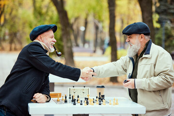 Portrait of two senior men playing chess in the park on a daytime in fall. Good game. Shaking...