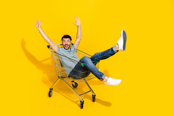 Full body photo of positive cheerful person sit market trolley raise hands isolated on yellow color...