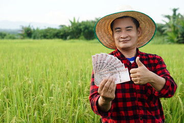 Asian farmer man is at paddy field, wears hat and red plaid shirt, hold Thai banknote money. ...