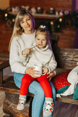 Obraz premium Portrait of a happy little girl sitting on her mother's lap and drinking hot chocolate on Christmas or New Year's Eve. Christmas mood, Christmas spirit, time together, motherhood, childhood