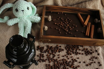 A soft toy lies on a table on a plush blanket. There is a wish card next to it. Coffee beans and a wooden tray complete the decor. Handmade. Hobby. Knitting. Gifts for the holidays.