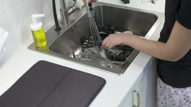 Woman washing with sponge forks knives spoons in kitchen sink under stream of water, female hands wash tableware closeup.