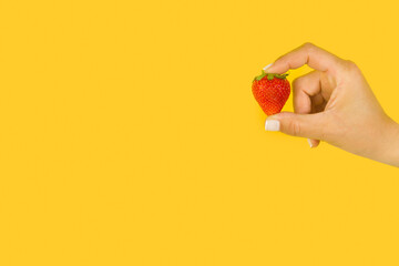 Mano de mujer sosteniendo una fresa fresca y natural sobre un fondo amarillo brillante liso y...