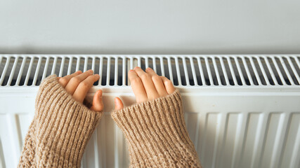 Child wearing brown woolly jacket warming cold hand in front of heating radiator in winter time. Electric or gas heater at home. Heating season, cold room, heating problems