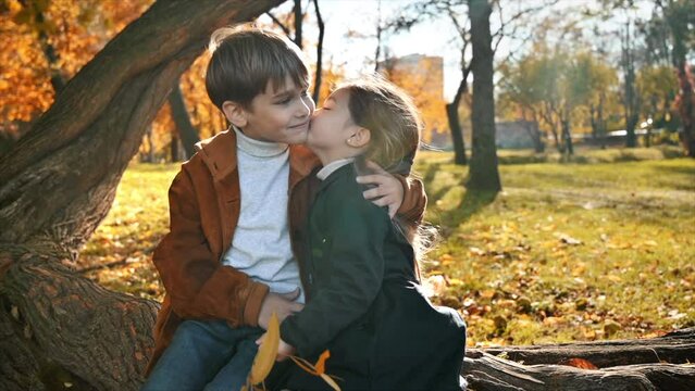 Happy family in an autumn park. Sister kisses her brother, he hugs her sitting on a tree trunk, yellowed trees around. Slow motion