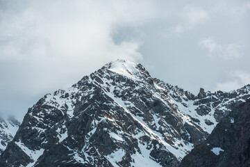 Obraz premium Dramatic landscape with sunlit high snowy mountain top in cloudy sky. White snow on black rocks in cloud sky. Awesome snow mountain peak in sunlight in changeable weather. Snowbound mountain in clouds