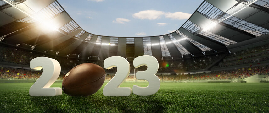 Leather american football ball on grass of football field between huge 3D numbers 2023 at stadium with spotlight. Concept of sport, art, energy, power