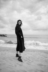 Young woman walking on sandy shore and enjoying view of sea in autumn. Stylish wear, skin care, brunette hipster enjoy the sea, happy model, dreamer. Monochrome