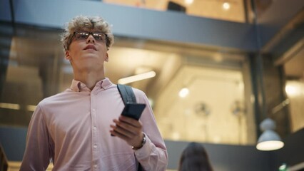 Portrait of University Student Checking his Text on his Smartphone and Smiling While he Walks Toward Classroom. Young White Man Male Crossing the Hall of a Big Building. Low Angle, Slow Motion Shot