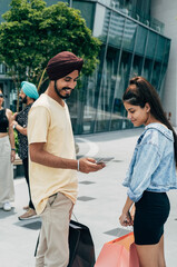 Happy Indian man using mobile phone while standing with teenage girl in front of the shopping mall with two unrecognizable multi-ethnic people who is talking in the background