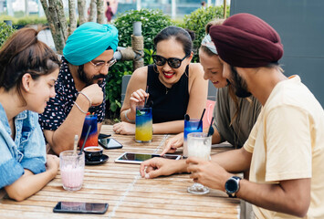 Happy friends watching something on a digital tablet and drinking cocktails and coffee while sitting together in the outdoor cafe