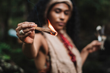 Imagen horizontal  de una mujer morena de cabello rizado al aire libre sosteniendo con su mano un trozo de palo santo con una llama de fuego mientras hace un ritual.   - obrazy, fototapety, plakaty