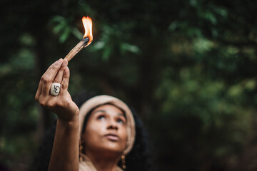 Imagen horizontal  de una mujer morena de cabello rizado al aire libre sosteniendo con su mano un trozo de palo santo con una llama de fuego mirando hacia el cielo mientras hace un ritual.   - obrazy, fototapety, plakaty