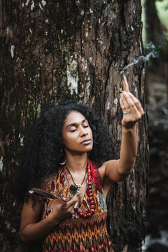 Imagen vertical de una mujer morena de cabello rizado recostada a un &aacute;rbol en el exterior haciendo un ritual mientras sostiene con su mano un trozo de palo santo encendido. 