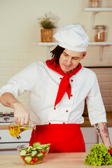 Young man with dreadlocks in uniform pouring olive oil on vegetable salad in kitchen. Positive chef preparing salad, adding healthy olive oil from jug.
