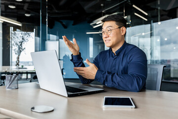 Successful Asian businessman talking on video call using laptop for remote communication with colleagues, boss in shirt smiling and gesturing with hands, online meeting with partners.