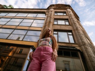 Sunny warm portrait of a cute blonde girl in a colorful top and pink pants on the street in summer. Summer vibe picture, beautiful woman is posing near the glass building.