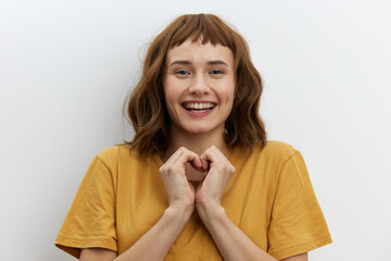 beautiful, attractive, redhead, romantic a woman poses standing in a yellow T-shirt on a white background and shows a heart sign with her fingers, smiling broadly and looking at the camera