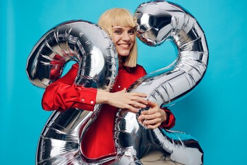 screaming loudly with delight, woman in a red shirt stands on a blue background and holds inflatable balloons in the shape of the number twenty-two in silver color, hugging them with her hands