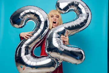 surprised, shocked woman in a red shirt stands with her mouth open on a blue background and holds inflatable balloons in the shape of the number twenty-two in silver color, hugging them with her hands