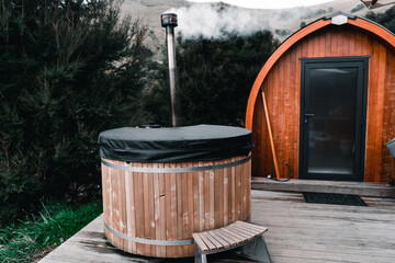 small round wooden jacuzzi with hot water and smoking fireplace outside of lonely small cozy wooden cabin far from civilization in the middle of the forest, te wepu pods akaroa, new zealand - obrazy, fototapety, plakaty