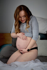Happy mother-to-be sits on the bed, feels kicks, caressing her big belly. A pregnant woman is talking to an unborn child.