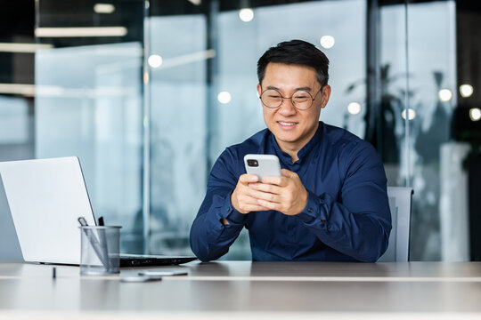 Successful Asian businessman working inside modern office building, man smiling and happy using smartphone to browse online pages, man in shirt and glasses with laptop.