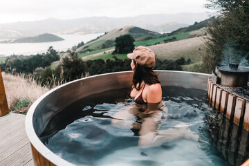 caucasian girl wearing swimsuit and brown hat inside round hot water jacuzzi looking at beautiful landscape of french farm bay calm sitting and relaxing near mountains and peaceful nature, te wepu - obrazy, fototapety, plakaty