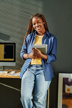 Young African American businesswoman in blue casualwear holding notebooks while standing by workplace with computer monitor