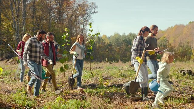Happy females and males with kids ready for volunteering project with seedlings of trees in forest. Outdoors. Environmental volunteers in wood going to plant new trees on nice fall day.