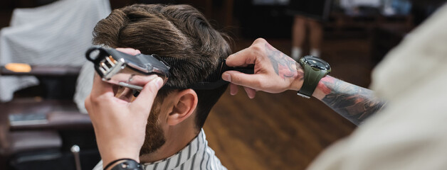 Tattooed barber combing and trimming hair of man in barbershop, banner.