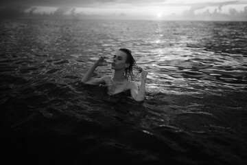 Black and white snapshot of a beautiful woman in the ocean. Dark portrait of a sensual gentle girl in the water in summer. Brunette woman is swimming and relaxing in the water before sunset. 