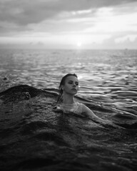 Black and white snapshot of a beautiful woman in the ocean. Dark portrait of a sensual gentle girl in the water in summer. Brunette woman is swimming and relaxing in the water before sunset. 