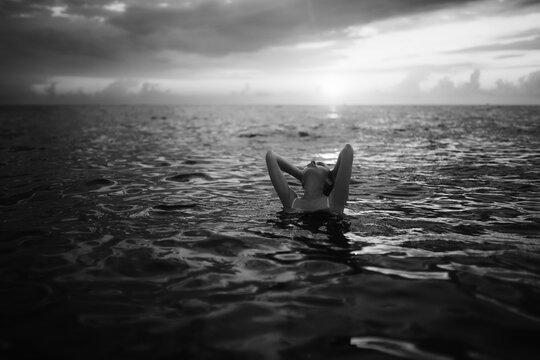 Black and white snapshot of a beautiful woman in the ocean. Dark portrait of a sensual gentle girl in the water in summer. Brunette woman is swimming and relaxing in the water before sunset.