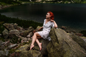 Elegant sexy lady with red hair in a white dress sits on the stone in the mountains and relaxes under sun rays. Fashion natural portrait as wallpaper, magazine design or advertising cover.