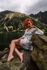 Elegant sexy lady with red hair in a white dress sits on the stone in the mountains and relaxes under sun rays. Fashion natural portrait as wallpaper, magazine design or advertising cover.