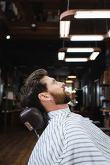 side view of bearded brunette man sitting in hairdressing cape in barbershop.