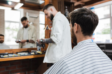 brunette man in hairdressing cape near blurred mirror and hairstylist in barbershop.