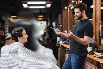 Positive teen boy in cape looking at hairstylist with scissors and comb in beauty salon.