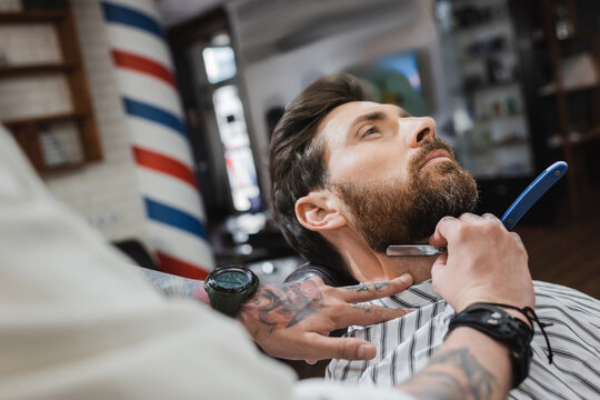 tattooed barber in wristwatch shaving client with straight razor.